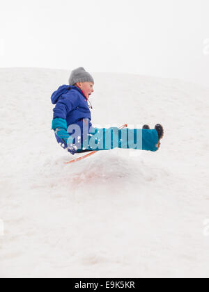 kleinen lachenden jungen schnell hinunter einen steilen Hügel in einer Wolke aus Schnee Rodeln Stockfoto