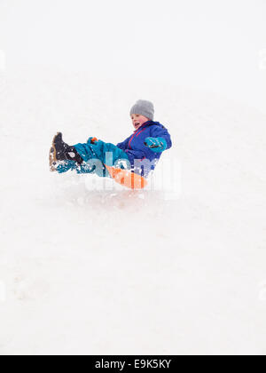 kleinen lachenden jungen schnell hinunter einen steilen Hügel in einer Wolke aus Schnee Rodeln Stockfoto