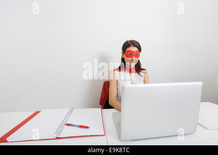 Junge Geschäftsfrau gekleidet als Superheld mit Laptop am Schreibtisch im Büro Stockfoto