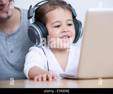 Kleines Mädchen mit Kopfhörern beim betrachten Laptop mit Vater am Tisch im Haus Stockfoto