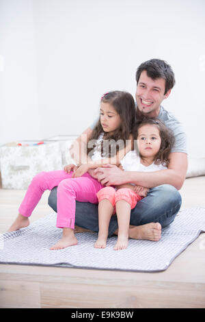 Lächelnder junge Mann mit Töchtern in seinem Schoß zu Hause sitzen Stockfoto