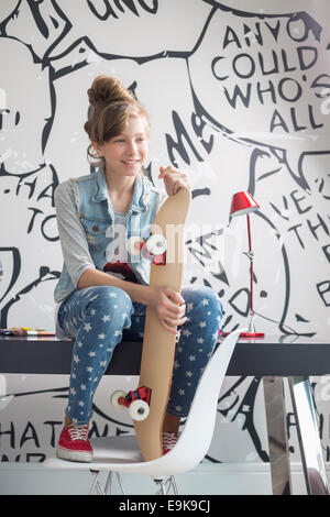 Mädchen mit Skateboard sitzt am Schreibtisch zu Hause in voller Länge Stockfoto