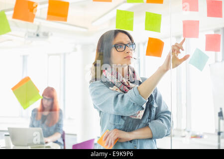 Kreative Geschäftsfrau Haftnotizen auf Glaswand mit Kollegen im Hintergrund im Büro arbeitet, lesen Stockfoto