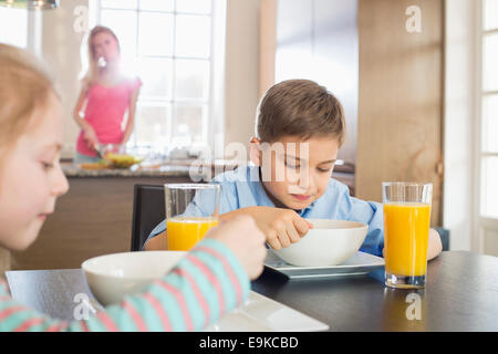 Geschwister mit Frühstück am Tisch mit Mutter, die Zubereitung von Speisen im Hintergrund Stockfoto