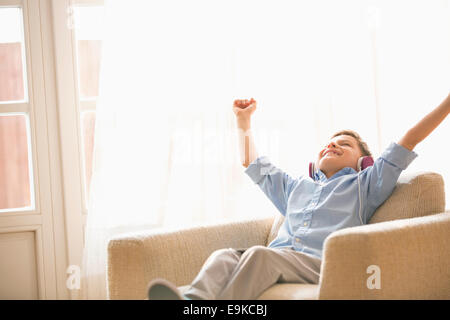 Fröhlicher Junge mit Armen angehoben genießen Musik während Sie gemütlich auf Sessel zu Hause Stockfoto