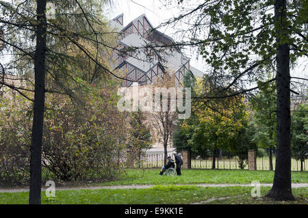 Krakau, Polen. 16. Oktober 2014. Eine Frau ist einen Rollstuhl mit im Hintergrund die Kirche Kosciol Zbawiciela (Kirche von unseres Erlösers) drängen. Nowa Huta, Krakau, Polen. Nowa Huta ist de Meest Oostelijke Voorstad van Krakau En de Thuisbasis van Een Groot Industrieel Komplex. Het Werd Ontworpen als Een Modell Communistische Stad En Gebouwd in de Sociaal-Realistische Stijl. Nowa Huta Werd Een Epi-Centrum van Het Verzet Tegen Het Communistische Bewind. Nowa Huta, Krakau, Polen. Nowa Huta ist die östlichste Vorort von Krakau und die Heimat einer großen Industriekomplex. Es wurde als Modell Kommunikation entwickelt. Stockfoto