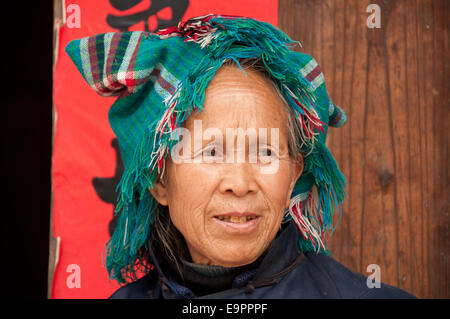 Die ältere Frau von Buyei (Bouyei) trägt ein gesäumtes traditionelles Kopftuch vor einem roten Banner und einer Holztür, Dorf Buyi, Provinz Guizhou, China Stockfoto