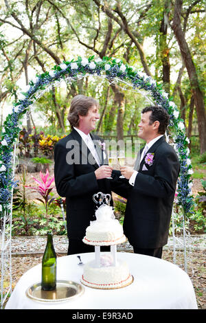 Glücklich schöne Homosexuell Paar Toasten mit Champagner unter Blumen Hochzeit Bogen. Hochzeitstorte mit zwei Pfleger im Vordergrund. Stockfoto