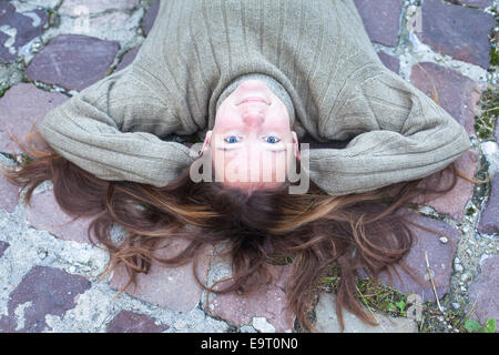 Junge schöne Mädchen in einen warmen Pullover liegend auf dem Kopfsteinpflaster, Ansicht von oben in die Kamera schauen. Stockfoto