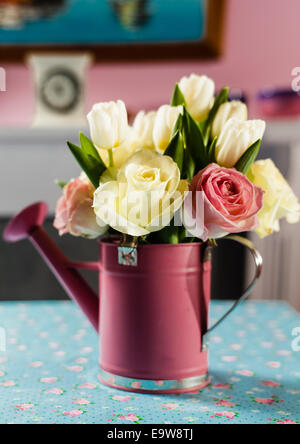 Rosa Creme Rosen weiße Tulpen rosa Gießkanne auf einem rosa und blauen Blumen Tisch Tuch Kaminsims und Uhr im Hintergrund Stockfoto