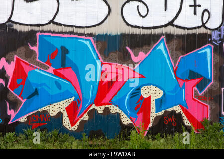 Abstrakte rosa und blau Graffiti über eine große braune gewellte Stahlwand mit Unkraut im Vordergrund. Stockfoto