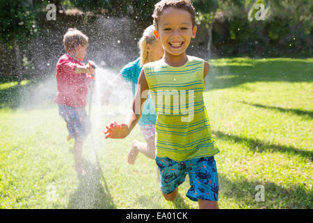 Drei Kinder jagten einander im Garten mit Wasser sprinkler Stockfoto