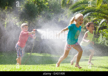 Drei Kinder im Garten jagen einander mit Wasser sprinkler Stockfoto
