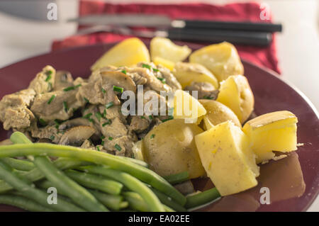 Schweinefleisch-Stroganoff, serviert auf einem roten Teller mit Kartoffeln und grünen Bohnen. Besteck im Hintergrund. Stockfoto