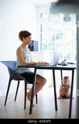 Mitte Erwachsene Mutter mit Kleinkind Tochter unter dem Tisch auf Laptop tippen Stockfoto