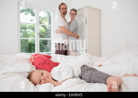 Babymädchen liegt auf Bett, Eltern im Hintergrund Stockfoto