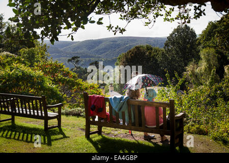 Zwei ältere Damen Schatten selbst Form der Sonne mit einem Regenschirm während sitzen und im Chat außerhalb auf einer Holzbank. Stockfoto