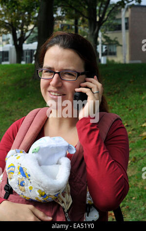 Eine lächelnde Mutter ein Baby zu tragen und mit Hilfe eines Mobiltelefons Stockfoto