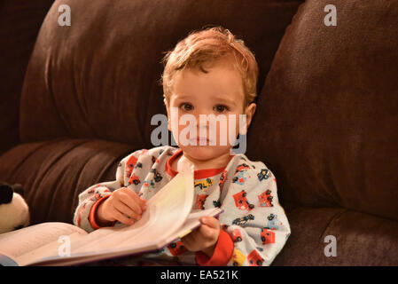 Ein kleiner Junge (zweieinhalb Jahre alt) im Schlafanzug saß auf dem Sofa ein Buch zu lesen, kurz vor dem Schlafengehen Stockfoto