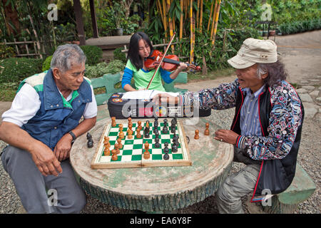 Zwei philippinische Männer spielen Schach, während ein junges Mädchen ihre Geige am Burnham Park, Baguio City auf den Philippinen spielt. Stockfoto