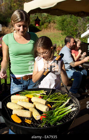 Tochter (8-9) holding Zangen und Mutter stehen vom Barbecue-grill Stockfoto