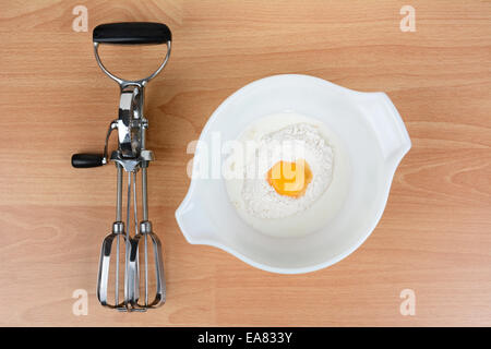 Hohen Winkel Schuss aus einer Schüssel mit Milch, Ei und Mehl mit einem alten altmodischen Handmixer auf dem Tisch daneben. Querformat. Stockfoto