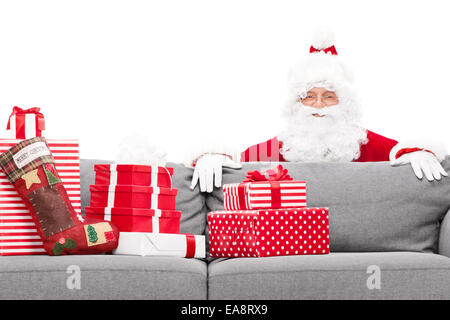 Santa Claus versteckt sich hinter einem Sofa voller Weihnachtsgeschenke auf weißen Hintergrund isoliert Stockfoto