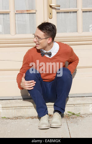 glücklich, gut aussehender Mann mit Brille und Pullover sitzen auf Stufen vor Haus und posiert beim Blick zur Seite, Vintage retro-ph Stockfoto