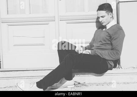 junger Mann in Pullover sitzen auf den Stufen vor Haus und posing, schwarz / weiß Foto Stockfoto