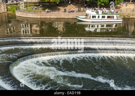 Touristenboot und Wasser fließt über / durch das Wehr am Fluss Avon in Bath, Somerset UK, in der Nähe von Pulteney Bridge. Stockfoto