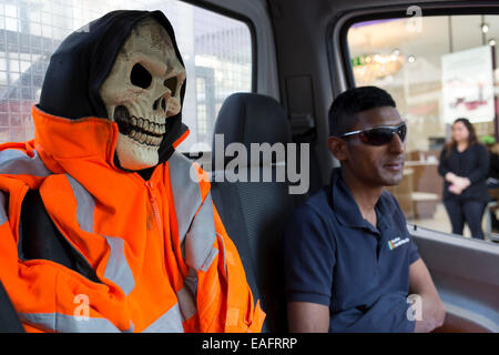 Mann in einem Kleinbus sitzt neben einem verkleideten Skelett auf dem Beifahrersitz. Halloween-Spaß mit einem Totenkopf in einem hohen viz Jacke. Londo Stockfoto