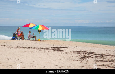 Sonnenanbeter sitzen unter bunten Sonnenschirmen an einem weißen Sandstrand Stockfoto