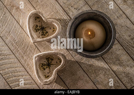 Zwei Herz-Dekorationen auf einem weißen gewaschenen Holztisch neben eine brennende Kerze in einen Steingut-Topf alles in niedrigen gerichtetes Licht. Stockfoto