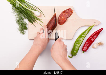 Weibliche Hände schneiden Scheiben rote Wurst auf ein Schneidbrett aus Holz umgeben mit verschiedenem Gemüse Stockfoto