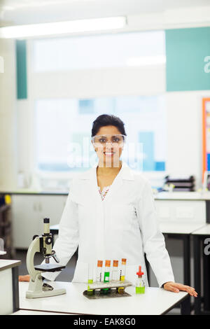 Porträt von lächelnden Lehrerin tragen von Schutzbrillen, stehend hinter Schreibtisch mit Mikroskop und Reagenzgläser im rack Stockfoto