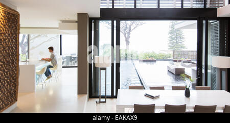 Junger Mann mit Laptop in moderne Küche, ein Essbereich mit Terrassentür und Pool im Vordergrund Stockfoto