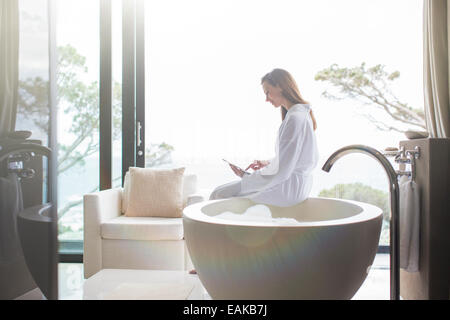 Frau mit weißen Bademantel sitzen am Rand des modernen Badewanne und mit digital-Tablette Stockfoto
