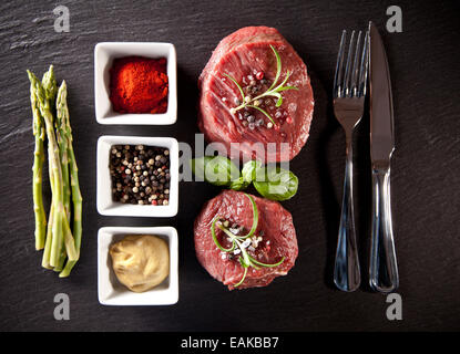 Stücke von roten rohes Fleisch Steaks mit Kräutern, serviert auf schwarzem Steinoberfläche. Erschossen von oben betrachtet. Stockfoto