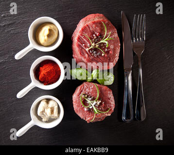 Stücke von roten rohes Fleisch Steaks mit Kräutern, serviert auf schwarzem Steinoberfläche. Erschossen von oben betrachtet. Stockfoto