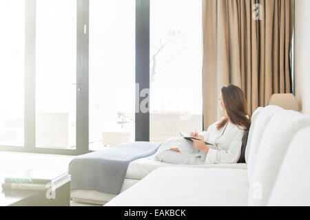 Frau trägt weißen Bademantel liegend auf dem Sofa im Wohnzimmer und Blick durch Fenster Stockfoto