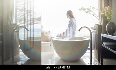 Frau trägt weißen Bademantel, sitzen am Rand der Badewanne und Blick durch Fenster Stockfoto