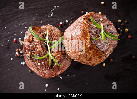 Stücke von rotem Fleisch Steaks mit Rosmarin auf schwarzen Steinoberfläche serviert. Aus oberen Geschossen Stockfoto