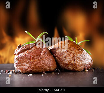 Stücke von rotem Fleisch Steaks mit Rosmarin auf schwarzen Steinoberfläche serviert. Feuer Flammen auf Hintergrund Unschärfe Stockfoto