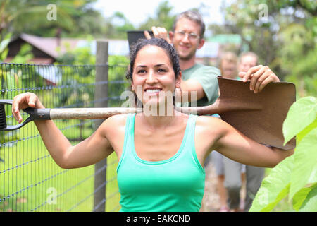 Lächelnde Frau tragen grüne Tank-Top mit Schaufel auf Schultern durch Gemeinschaftsgarten Stockfoto