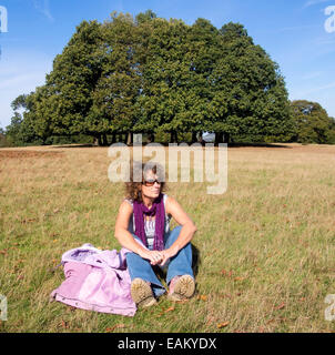 Entspannte junge Frau saß auf dem Rasen in einem Feld in der Nähe von einigen Bäumen Stockfoto