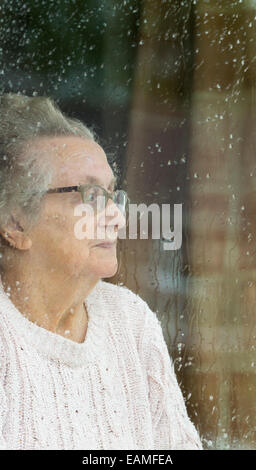 Neunzigjährige Frau, die an einem regnerischen Tag aus dem Fenster schaut. Selbstisolation, Quarantäne, Coronavirus, Konzept der sozialen Distanzierung... Stockfoto