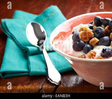 Joghurt mit frischen Heidelbeeren in einer Schüssel orange Frühstück mit einem silbernen Löffel auf eine türkisfarbene Serviette auf einem antiken Holztisch Stockfoto
