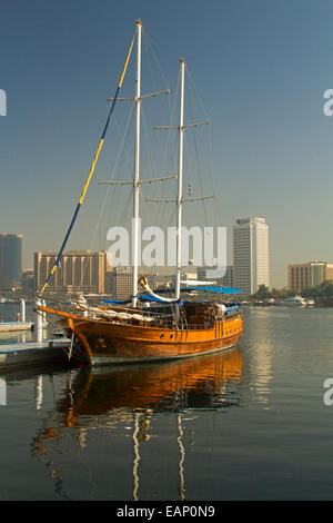 Hölzerne Yacht ankern & spiegelt sich in ruhigen blauen Wasser des Dubai Creek mit modernen Wolkenkratzern der Stadt Dubai im Hintergrund unter blauem Himmel Stockfoto