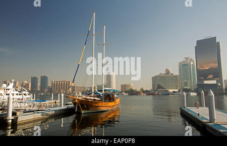 Hölzerne Yacht ankern & spiegelt sich in ruhigen blauen Wasser des Dubai Creek mit Glas ummauerten Wolkenkratzer & anderen modernen Gebäuden der Stadt von Dubai in der Nähe Stockfoto