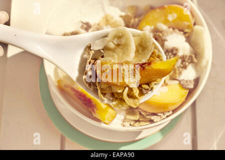 Frühstück in der Sonne, Müslischale, Pfirsich und Banane, Joghurt auf Pastellblau Platte. Weiß auf Holz Stockfoto